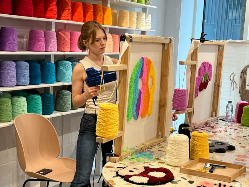 A woman stands at a table using a tufting gun to create a rainbow coloured design on fabric